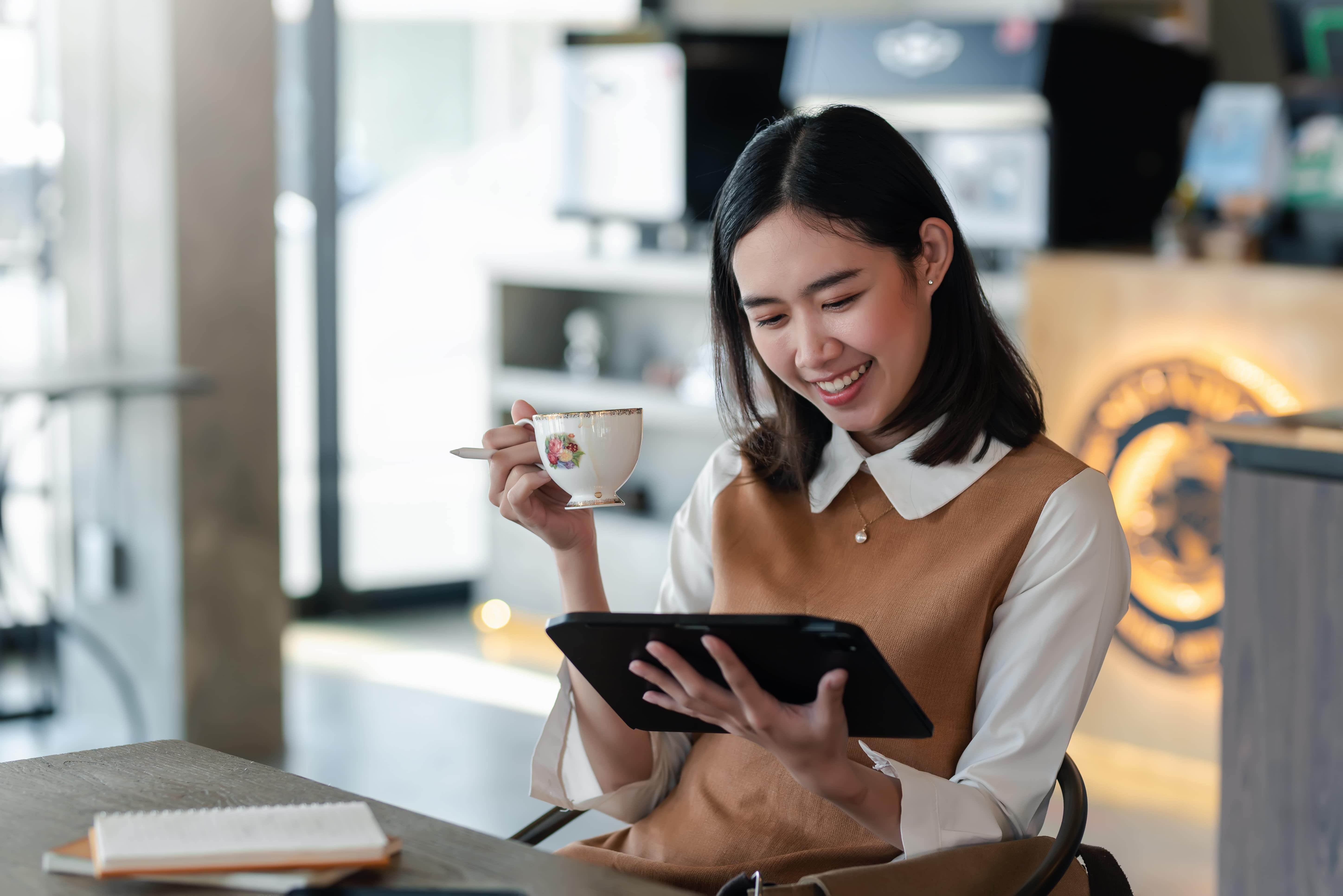 Une femme assise dans un café lit sur une tablette et tient une tasse de thé.