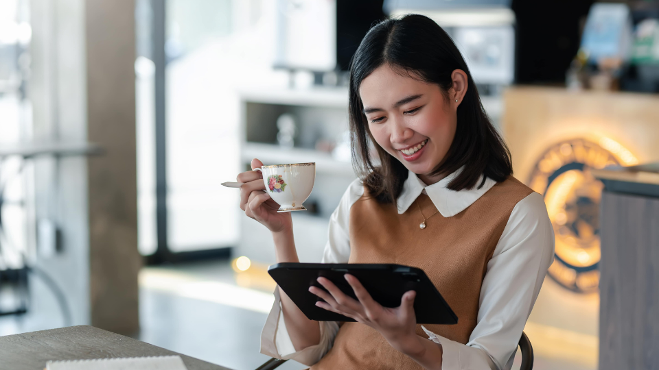 Une femme assise dans un café lit sur une tablette et tient une tasse de thé.