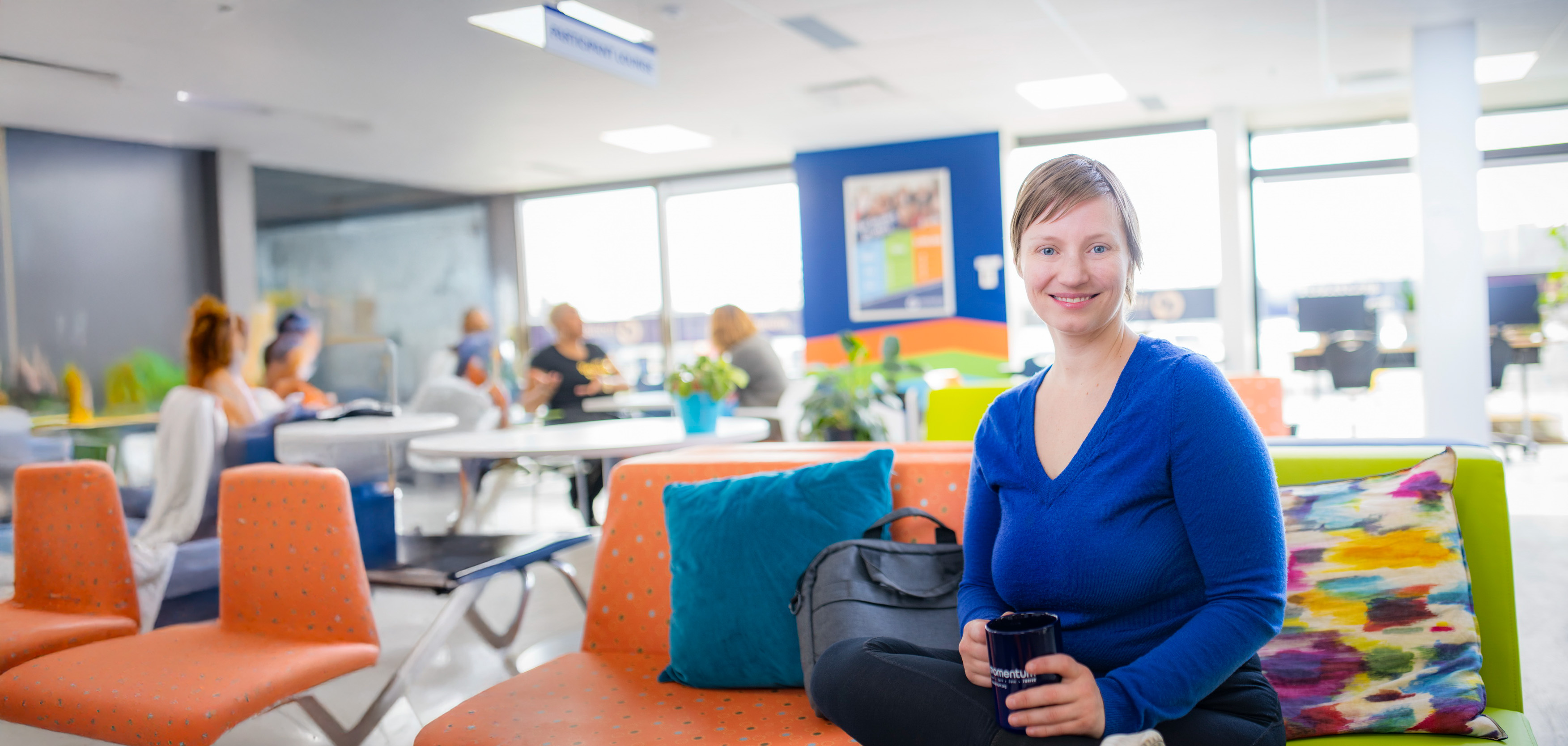 Une femme est assise, jambes croisées, sur un fauteuil dans un bureau, tandis que d’autres personnes sont assises à des tables en arrière-plan. La femme sourit à la caméra et tient une tasse.